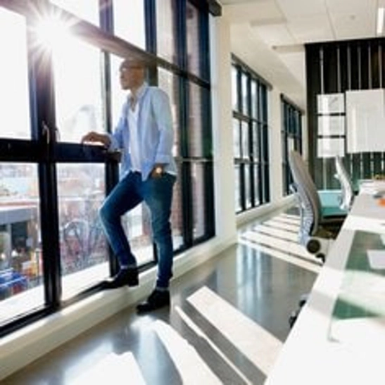 man in open blue button down white tshirt and jeans looking out windows of business building